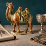 Tang-inspired art still life with a glazed camel, ink horse scroll, silver cup, and patterned silk